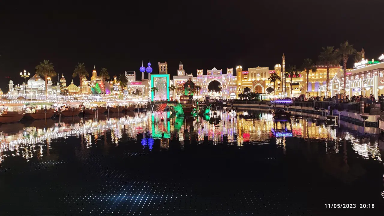 บูธและแสงไฟสะท้อนผิวน้ำที่ Global Village ยามค่ำ ภาพจาก Wikimedia Commons ผู้ถ่าย Philosographer สัญญา CC BY-SA 4.0