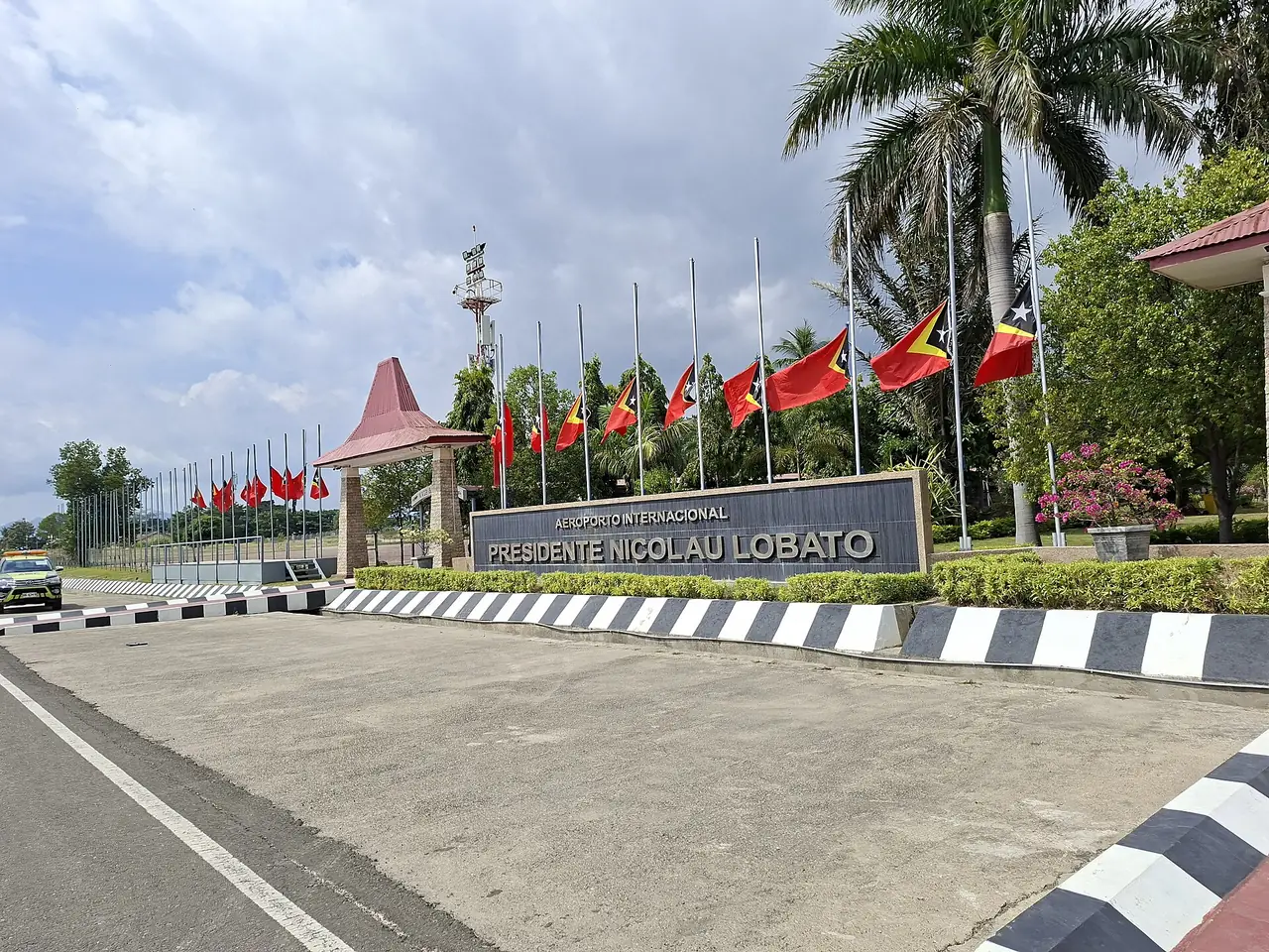 ป้ายหน้าสนามบินดิลีและธงชาติติมอร์-เลสเตครึ่งเสา ภาพจาก Wikimedia Commons ผู้ถ่าย Swarabakti สัญญา CC BY 4.0 ถ่ายเมื่อ 26 มิ.ย. 2569 ตามชื่อไฟล์