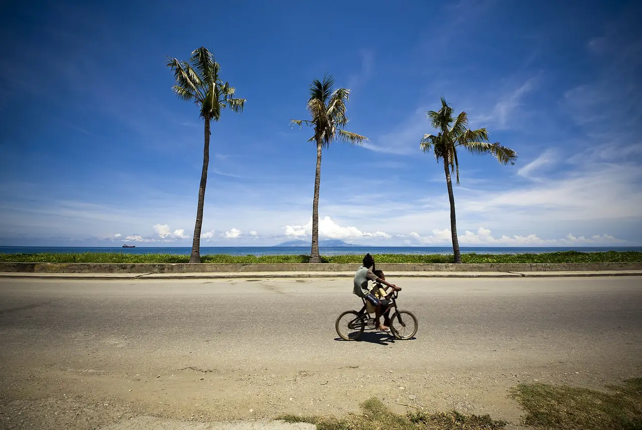 ถนนริมหาดในดิลีมีต้นมะพร้าวและคนปั่นจักรยาน ภาพจาก Wikimedia Commons ผู้ถ่าย Thomas Halloran สัญญาสาธารณสมบัติ ภาพเก่า