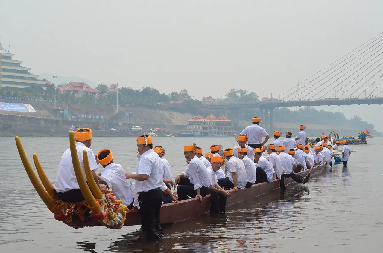 ลูกเรือสวมผ้าโพกส้มนั่งเรือมังกรยาวบนแม่น้ำ มีสะพานขึงอยู่ด้านหลัง ภาพจาก Wikimedia Commons ผู้ถ่าย Peter Olshevsky สัญญา CC BY-SA 4.0 ชื่อไฟล์เขียนว่า Water-Splashing Festival แต่ภาพเห็นเรือมังกร ไม่เห็นการสาดน้ำ และไม่ระบุปีถ่าย