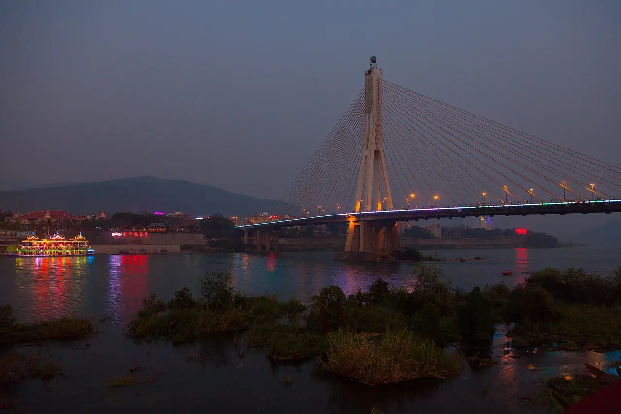 สะพานขึงข้ามแม่น้ำล้านช้างที่จิ่งหงยามค่ำ มีเรือประดับไฟทางซ้าย ภาพจาก Wikimedia Commons ผู้ถ่าย Chlukoe สัญญา CC BY-SA 4.0 ไม่ระบุปีถ่าย