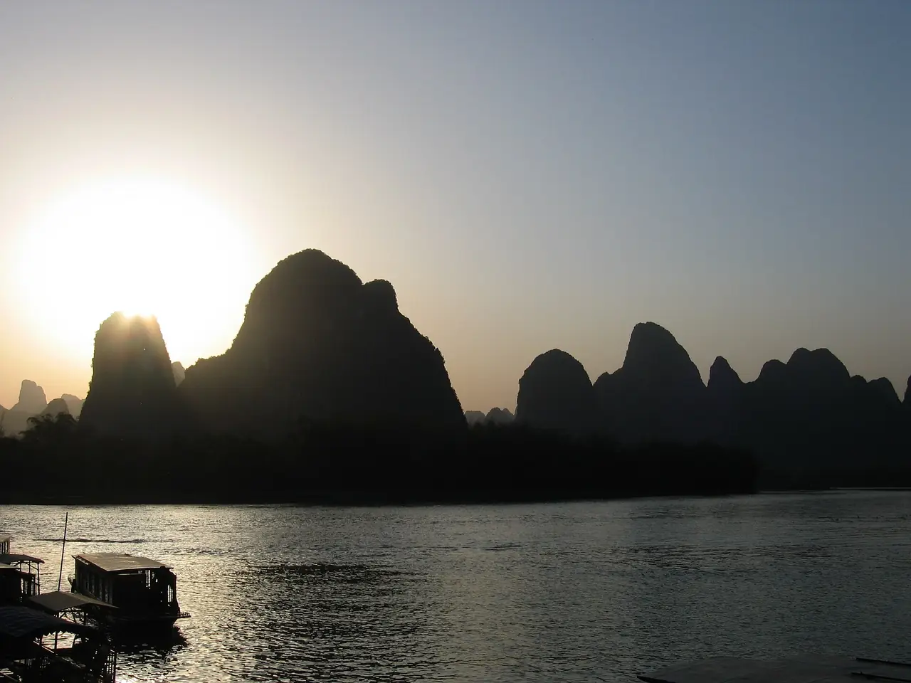 เงาเขาหินปูนตัดกับแสงอาทิตย์ตก มีเรือจอดริมแม่น้ำหลี่ ภาพจาก Wikimedia Commons ผู้ถ่าย Chinatravelsavvy สัญญา CC BY-SA 3.0 ชื่อไฟล์ระบุว่าเป็นแม่น้ำหลี่ที่ซิงผิง ไม่ระบุปีถ่าย
