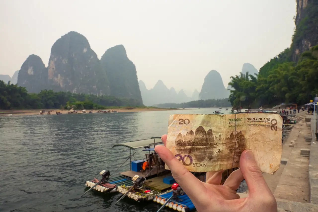 ผู้ถ่ายถือธนบัตร 20 หยวนทาบกับฉากแม่น้ำหลี่ที่ซิงผิง มีเรือแพเครื่องยนต์จอดอยู่และเขาหินปูนเรียงเป็นฉากหลัง ภาพจาก Wikimedia Commons ผู้ถ่าย Michaeljscott93 สัญญา CC BY-SA 4.0 ไม่ระบุปีถ่าย