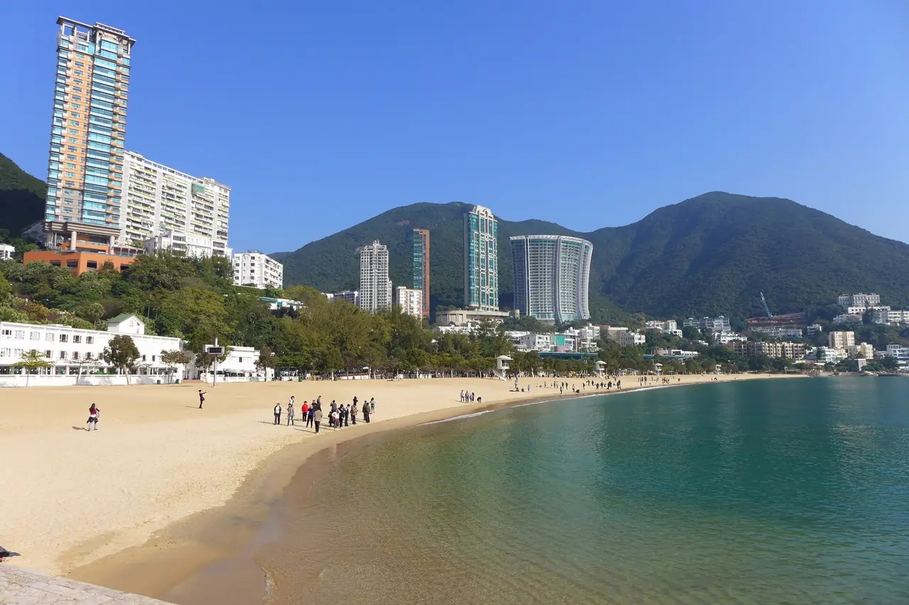 หาด Repulse Bay มองไปทางเหนือ เห็นตึกริมหาดและเนินเขา ภาพถ่ายปี 2558 อาจมีรายละเอียดอาคารต่างจากปัจจุบัน ภาพจาก Wikimedia Commons ผู้ถ่าย Wing1990hk สัญญา CC BY 3.0