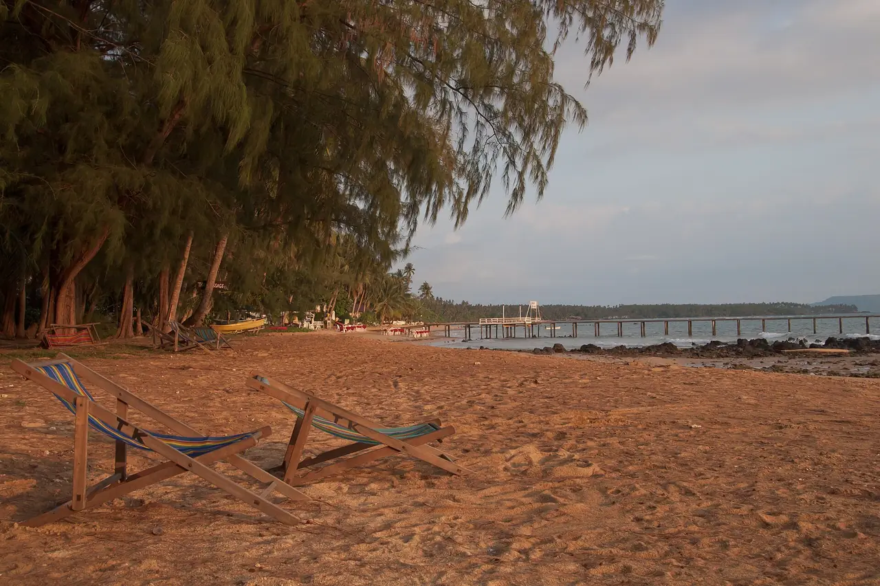 หาดทรายสีส้มและต้นสนริมหาด มีเก้าอี้พับสองตัวและสะพานไม้ไกล ๆ บนเกาะหมาก ภาพของ Vyacheslav Argenberg จาก Wikimedia Commons ไม่ระบุชื่ออ่าว