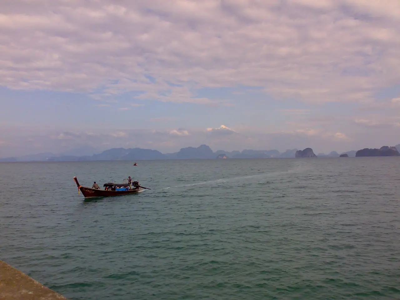 เรือหางยาวไม้สีแดงแล่นกลางน้ำสงบ เห็นภูเขาหินปูนไกล ๆ ของอ่าวพังงา ภาพของ Jaakko H. จาก Wikimedia Commons ตามคำบรรยายภาพบน Commons เป็นเรือหางยาวที่กำลังเข้าใกล้เกาะยาวน้อย
