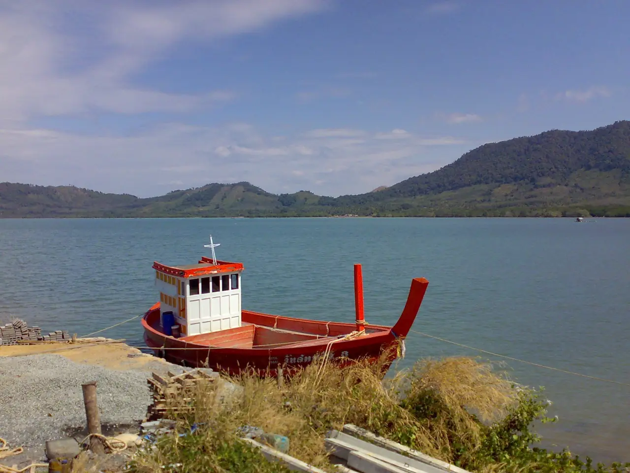เรือประมงไม้สีแดงจอดริมน้ำ มีเทือกเขาและทะเลเป็นฉากหลัง ภาพของ Jaakko H. จาก Wikimedia Commons ตามคำบรรยายภาพบน Commons ถ่ายที่ศูนย์วิจัยทางทะเลบนเกาะยาวใหญ่ ภาพเก่า ไม่ได้ระบุปี