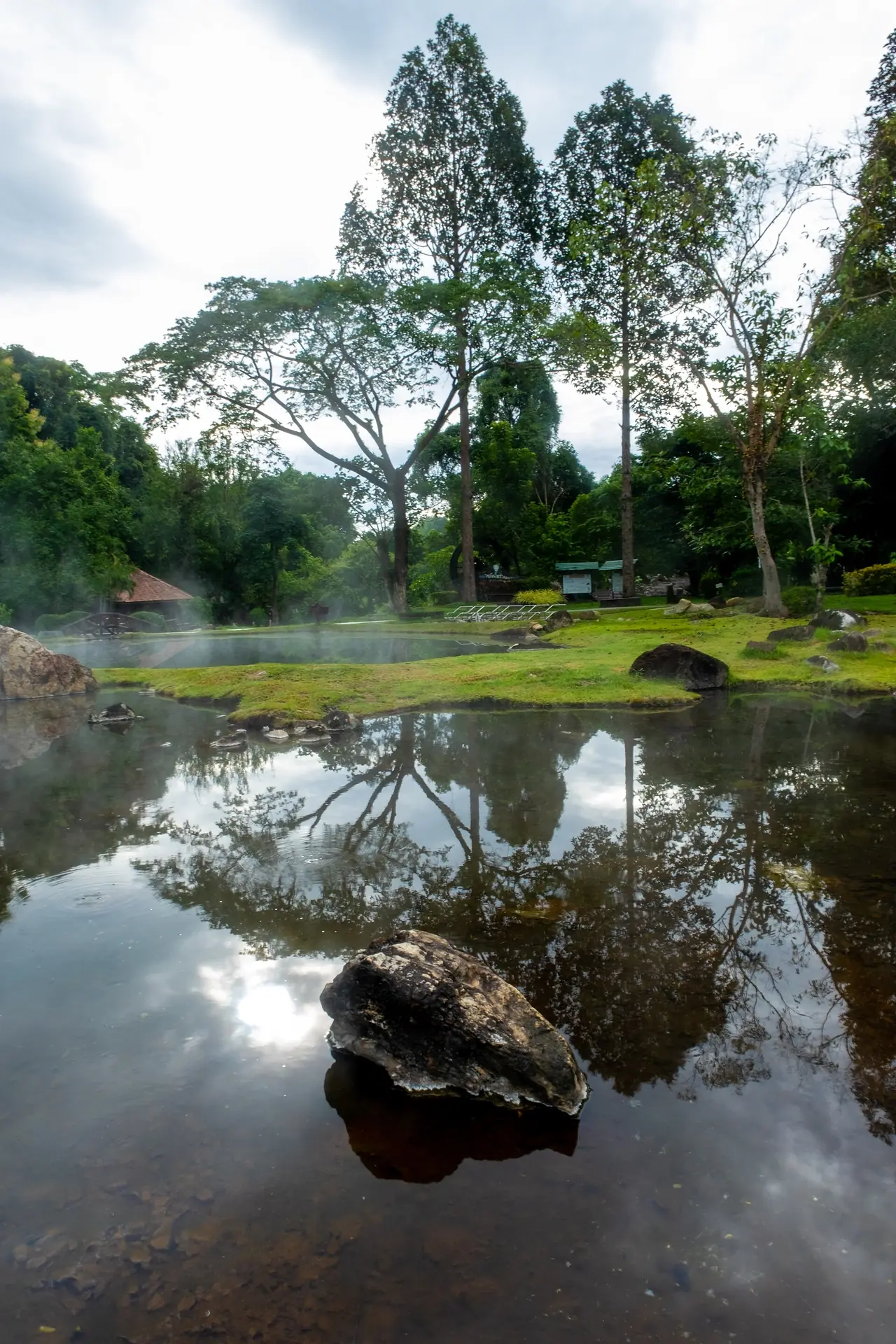 ผืนน้ำนิ่งสะท้อนต้นไม้และก้อนหินที่บ่อน้ำพุร้อน ภาพของ Jomis 9 จาก Wikimedia Commons ใบอนุญาต CC BY-SA 4.0 ถ่ายปี 2568 ภาพใช้เป็นภาพประกอบบรรยากาศ ไม่ใช่การยืนยันว่าบ่อไหนลงอาบได้
