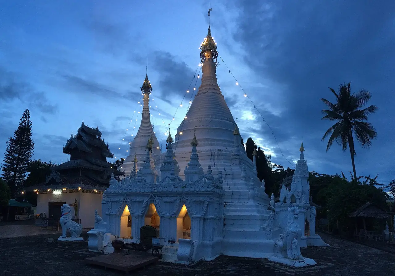 พระเจดีย์สีขาวที่ดอยกองมูยามพลบค่ำ มีไฟประดับสายและวิหารไม้ด้านซ้าย ภาพถ่ายจาก Wikimedia Commons