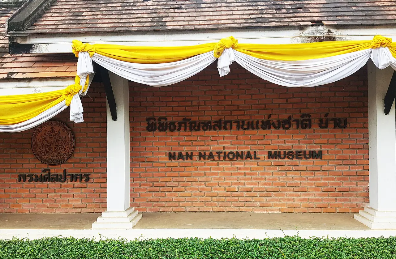 ป้ายชื่อพิพิธภัณฑสถานแห่งชาติ น่าน ติดบนผนังอิฐ มีตรากรมศิลปากรอยู่ด้านซ้าย ภาพถ่ายจาก Wikimedia Commons
