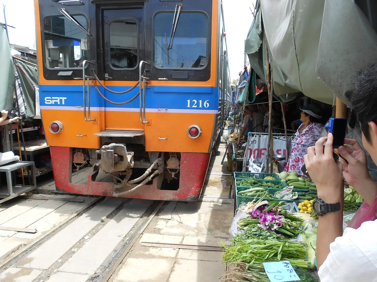รถไฟสายแม่กลองผ่านตลาดมหาชัย ภาพของ Raki_Man จาก Wikimedia Commons ใบอนุญาต CC BY 3.0 ชื่อไฟล์ระบุว่าเป็นตลาดมหาชัยแม่กลอง ไม่ระบุปีที่ถ่าย