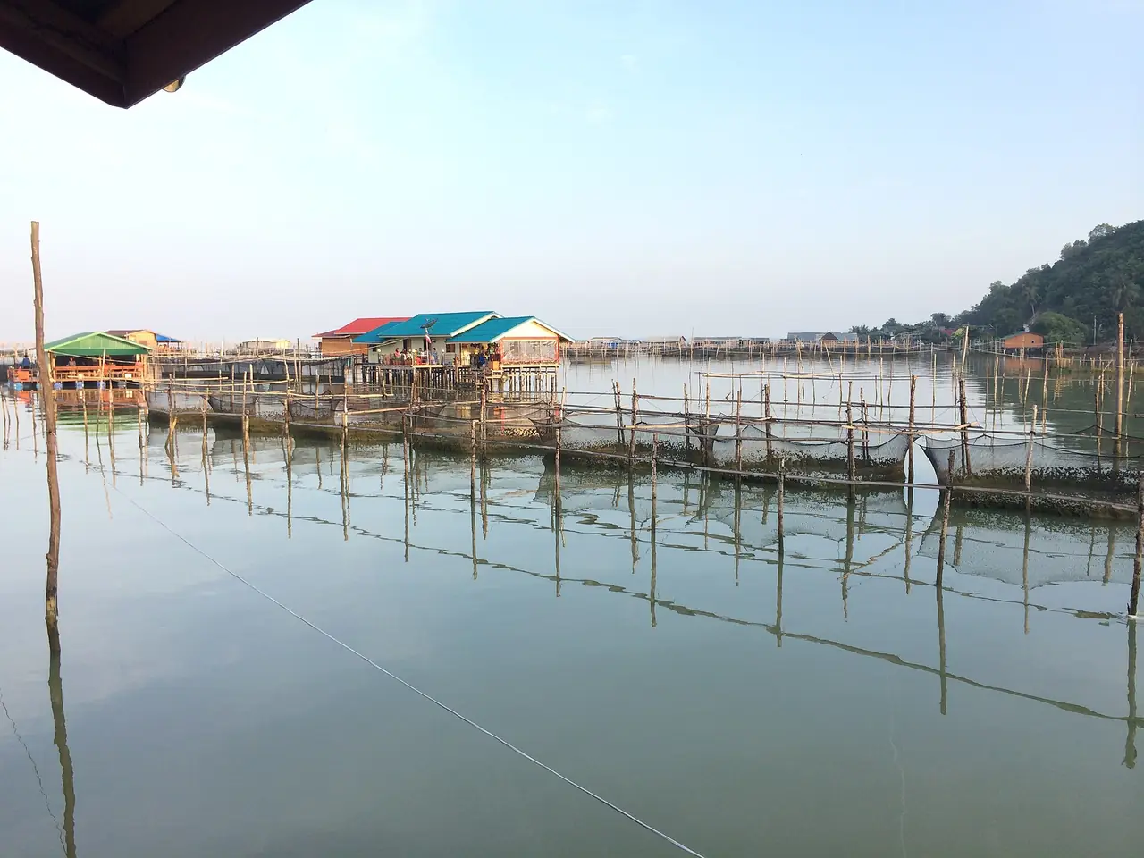 เสาไม้กระชังปลาและกระท่อมบนผิวน้ำนิ่งของทะเลสาบสงขลา มองจากเกาะยอ