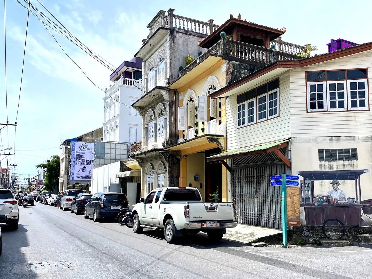 ถนนในย่านเมืองเก่าสงขลาที่มีตึกแถวสีเหลืองและรถเข็นกาแฟบนผนังด้านขวา ภาพของ Slleong จาก Wikimedia Commons (CC0)