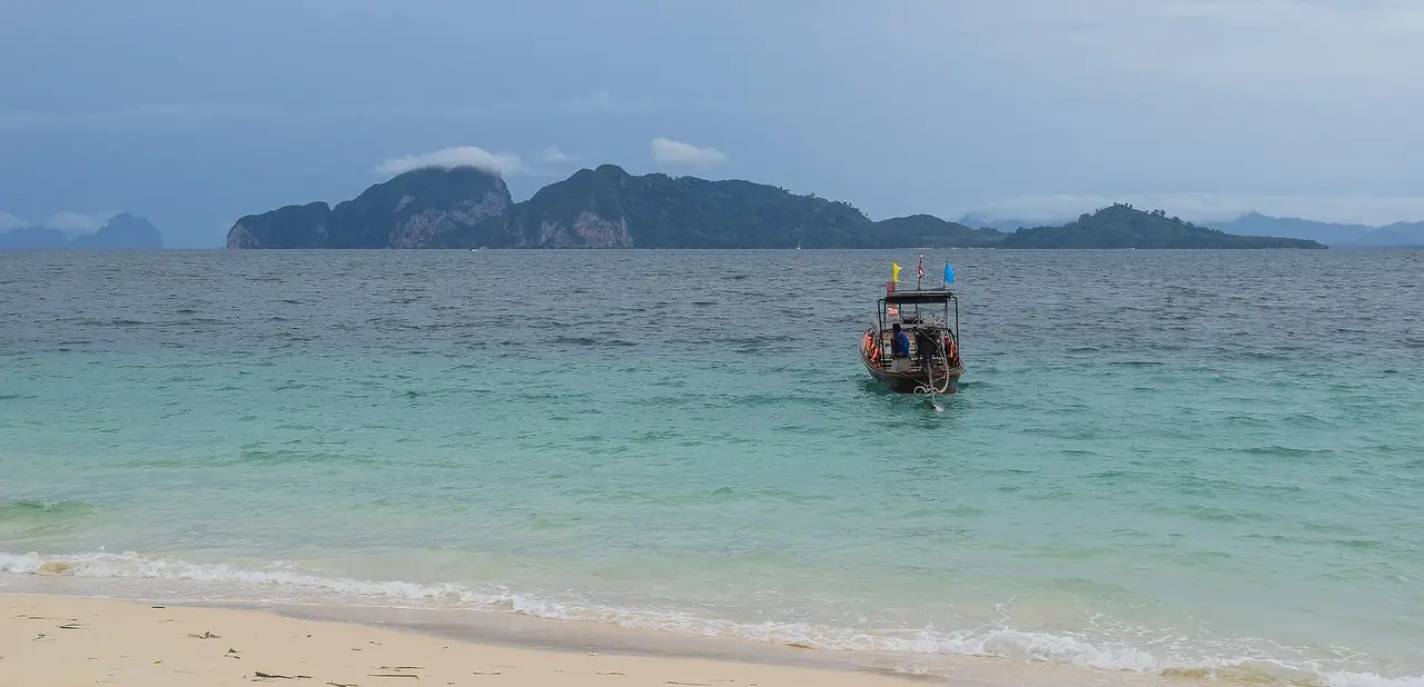 เกาะมุกมองจากชายหาดฝั่งตรงข้าม มีเรือหางยาวจอดอยู่ใกล้ฝั่ง ภาพถ่ายจาก Wikimedia Commons