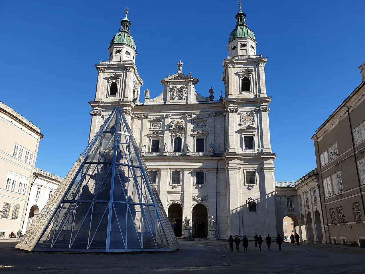 หน้าอาสนวิหารซาลซ์บูร์กและลาน Domplatz เห็นปิรามิดกระจกที่ตั้งอยู่หน้าโบสถ์ ภาพจาก Wikimedia Commons ผู้ถ่าย Strubbl สัญญา CC BY-SA 4.0 (ถ่ายเดือนกุมภาพันธ์ 2565)
