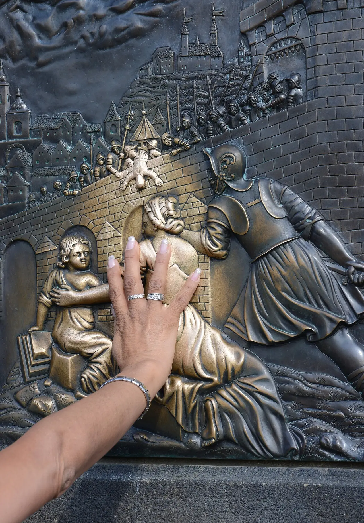 มือลูบแผ่นโลหะนูนต่ำที่ฐานรูปปั้นบนสะพานชาร์ลส์ ธรรมเนียมที่ CzechTourism เล่าว่าลูบแล้วได้โชค ภาพจาก Wikimedia Commons ผู้ถ่าย Alvesgaspar สัญญา CC BY-SA 4.0