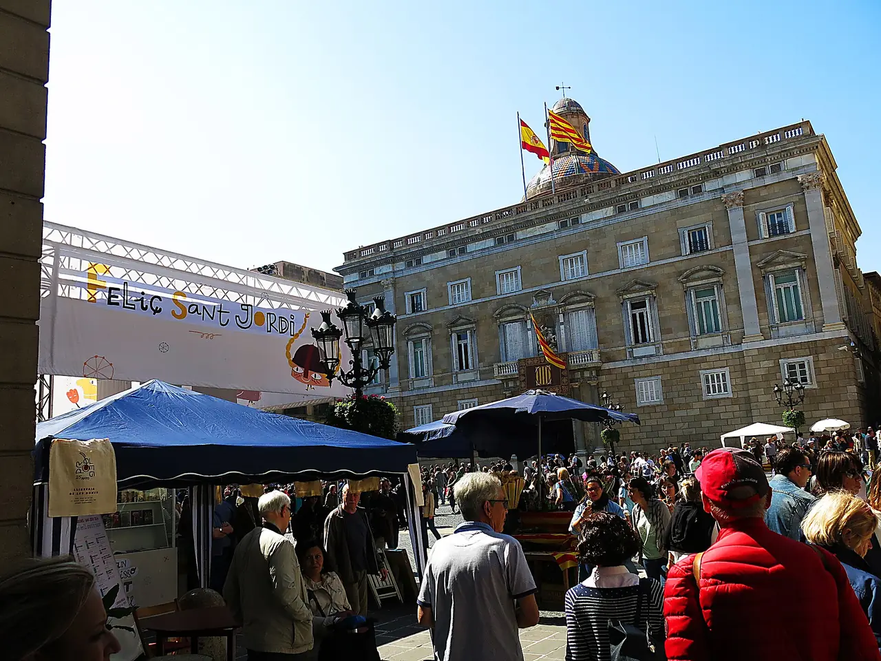 Plaça Sant Jaume ในวัน Sant Jordi ปี 2016 มีผู้คนและแผงจัดงาน ด้านหลังเป็น Palau de la Generalitat ภาพถ่ายจาก Wikimedia Commons ผู้ถ่าย Enric สัญญา CC BY-SA 4.0 ภาพช่วงงานพิเศษ บรรยากาศปกติต่างไป