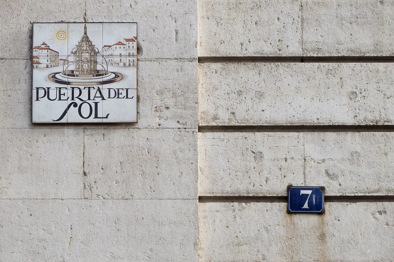 ป้ายกระเบื้องชื่อ Puerta del Sol บนผนังอาคารในมาดริด ภาพถ่ายจาก Wikimedia Commons ผู้ถ่าย Carlos Delgado สัญญา CC BY-SA 3.0 เป็นภาพป้ายถนน ไม่ใช่ภาพจัตุรัสทั้งหมด