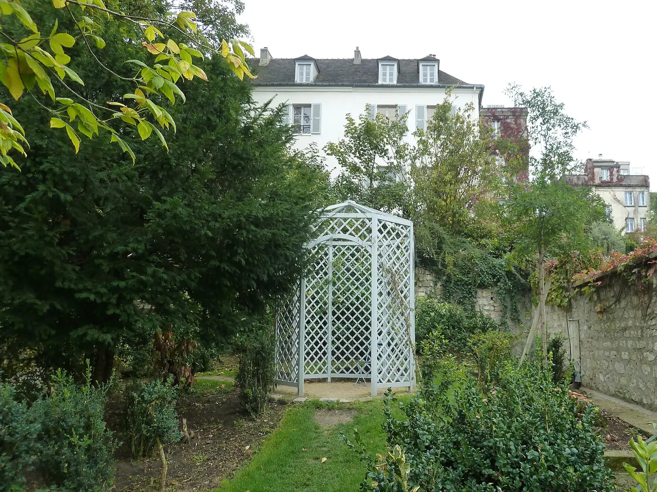สวน Renoir ของ Musée de Montmartre มีซุ้มสีขาวกลางสวนและบ้านอยู่ด้านหลัง ภาพจาก Wikimedia Commons ผู้ถ่าย Pierre André Leclercq สัญญา CC BY-SA 4.0