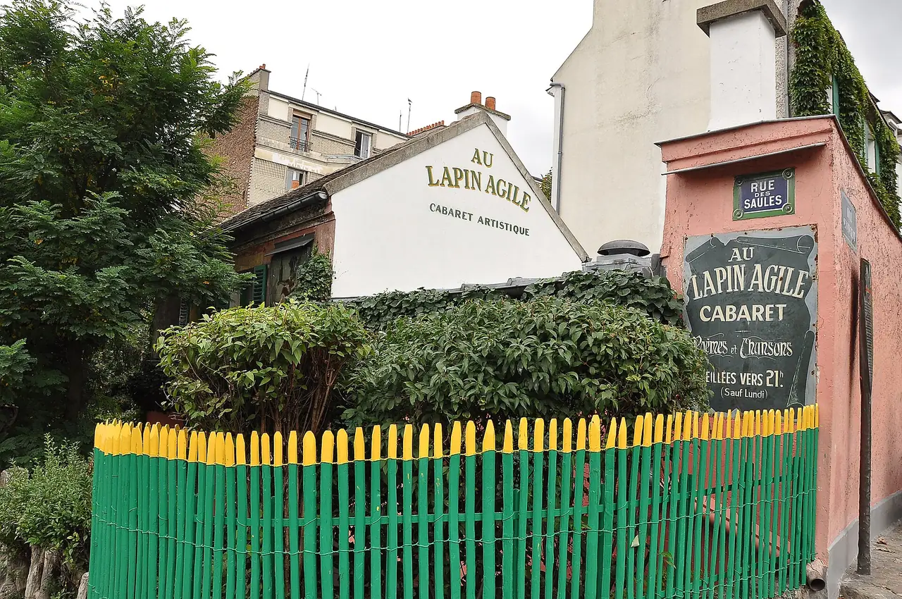ร้านคาบาเรต์ Au Lapin Agile ที่รั้วสีเขียวเหลืองและป้ายถนน Rue des Saules ในมงมาร์ต ภาพจาก Wikimedia Commons ผู้ถ่าย Moonik สัญญา CC BY-SA 3.0