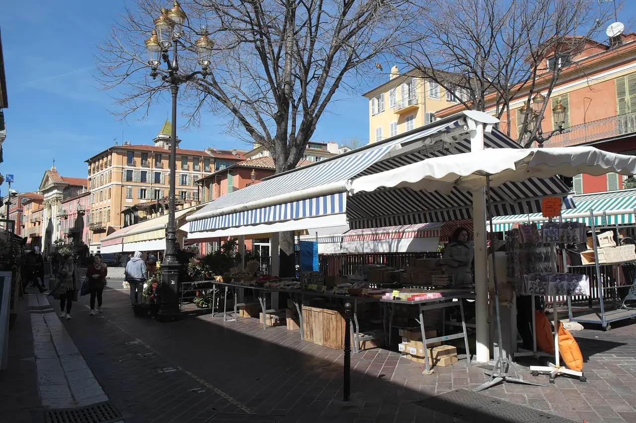 ตลาด Cours Saleya ในเมืองเก่านีซ แผงขายของใต้ร่มขาวและอาคารสีส้มสองฝั่ง ภาพจาก Wikimedia Commons ผู้ถ่าย GFreihalter สัญญา CC BY-SA 4.0