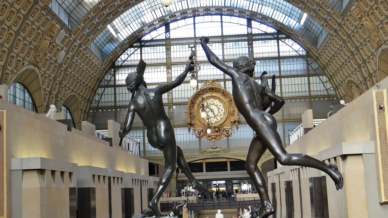 โถงใหญ่ของ Musée d'Orsay ที่มีนาฬิกาทองอยู่กลางหลังคาโค้งกระจกและรูปปั้นสัมฤทธิ์สองตัวด้านหน้า ภาพจาก Wikimedia Commons ผู้ถ่าย Ibex73 สัญญา CC BY-SA 4.0