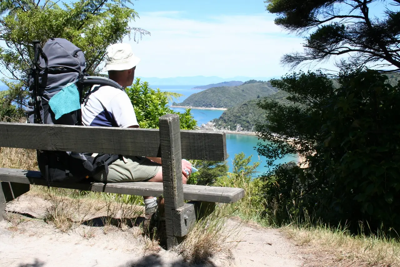 นักเดินป่านั่งพักบนม้านั่งที่จุดชมวิวบน Abel Tasman Coast Track มองลงอ่าวสีฟ้า ภาพจาก Wikimedia Commons ผู้ถ่าย Department of Conservation สัญญา CC BY 2.0 ไม่ระบุปี