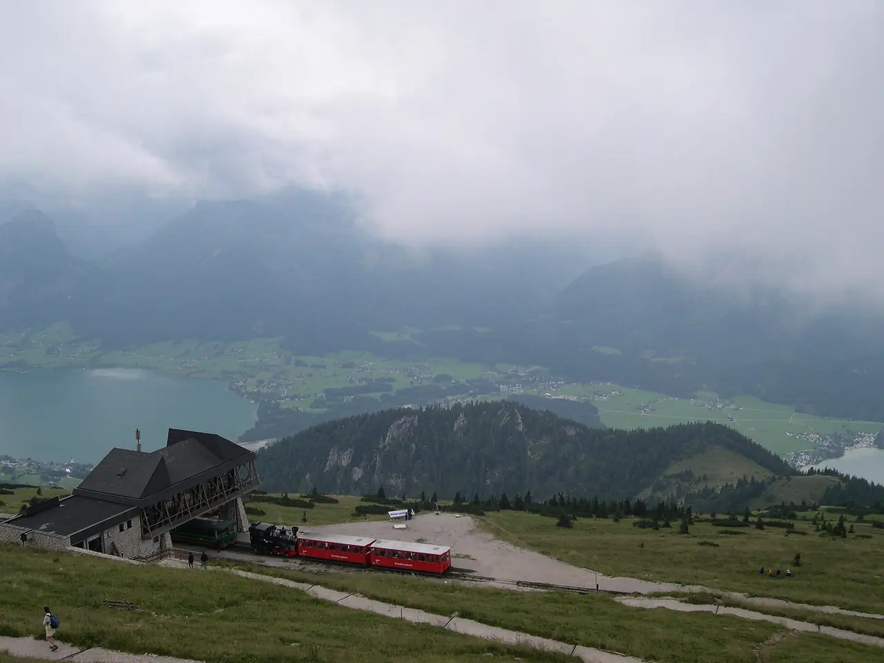สถานียอดของ Schafbergbahn และทะเลสาบด้านล่างจากยอด Schafberg ภาพจาก Wikimedia Commons ผู้ถ่าย malouette สัญญา CC BY-SA 2.0 ถ่ายวันที่มีเมฆ ปีถ่ายไม่ระบุในที่เราตรวจ