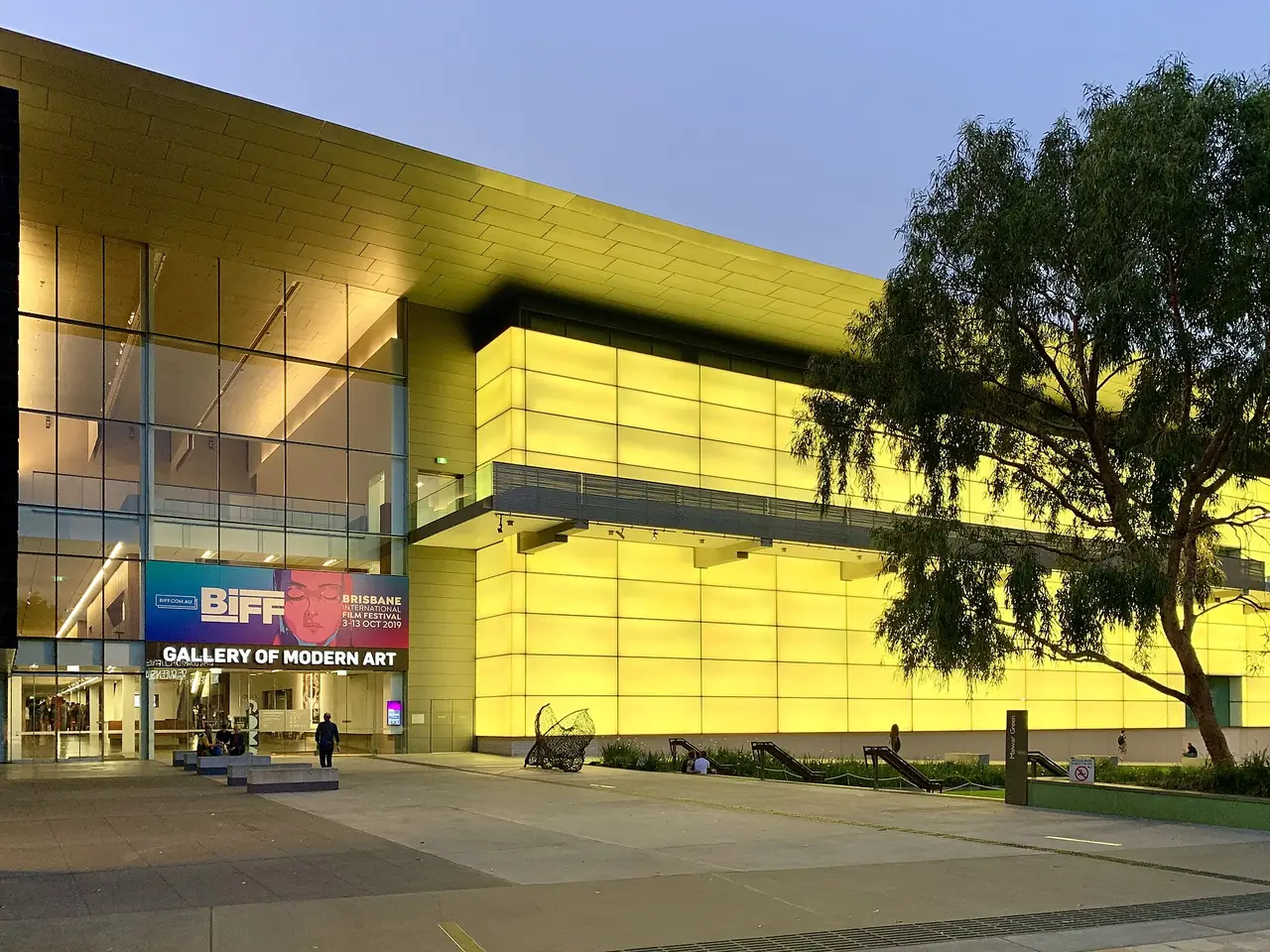 อาคาร Gallery of Modern Art (GOMA) ยามพลบค่ำ ริมแม่น้ำบริสเบน ป้ายหน้าอาคารเป็นของเทศกาลภาพยนตร์ปี 2562 ภาพจาก Wikimedia Commons ผู้ถ่าย Chris Olszewski สัญญา CC BY-SA 4.0
