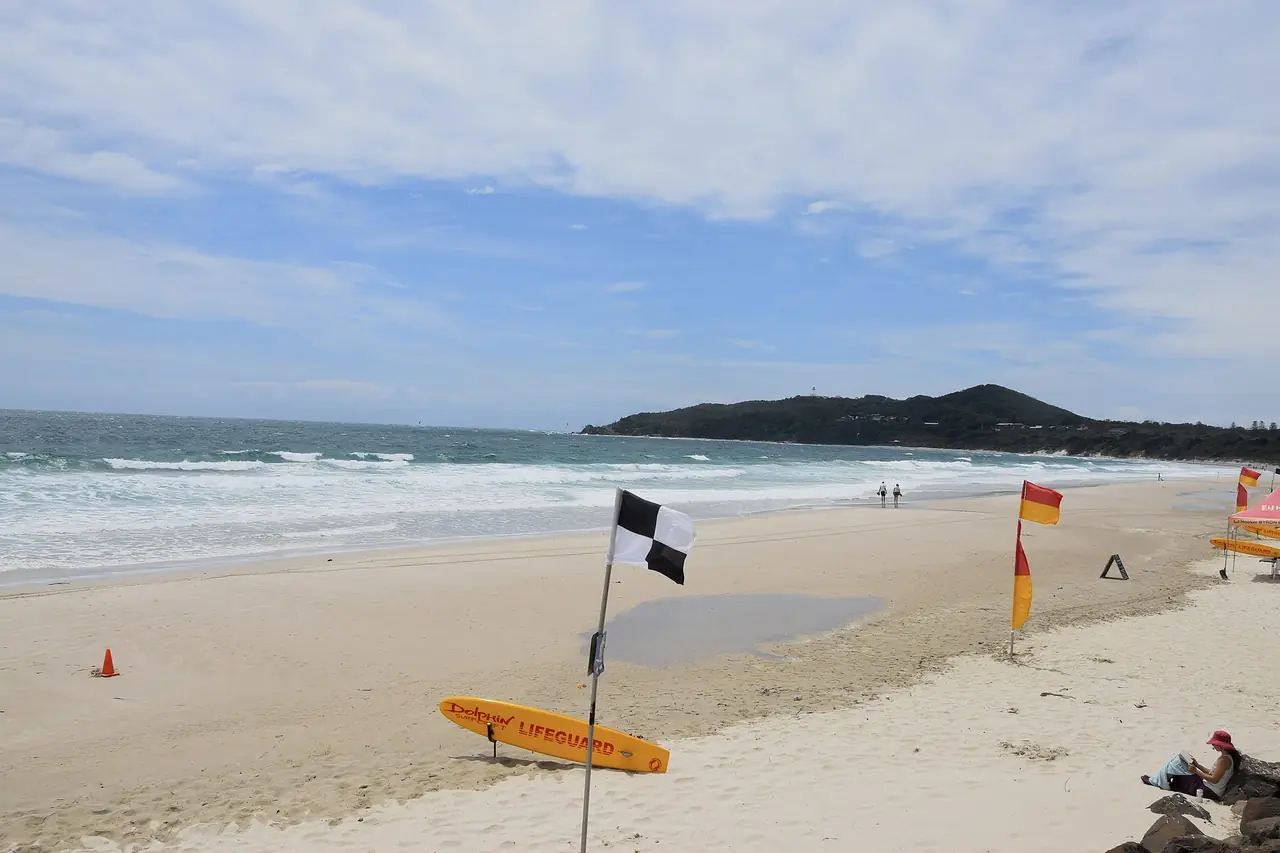 หาด Main Beach ที่ Byron Bay มีธงเหลืองแดงและเรือกระดานของไลฟ์การ์ด และแหลม Cape Byron เป็นฉากหลัง ภาพจาก Wikimedia Commons ผู้ถ่าย Ank Kumar สัญญา CC BY-SA 4.0