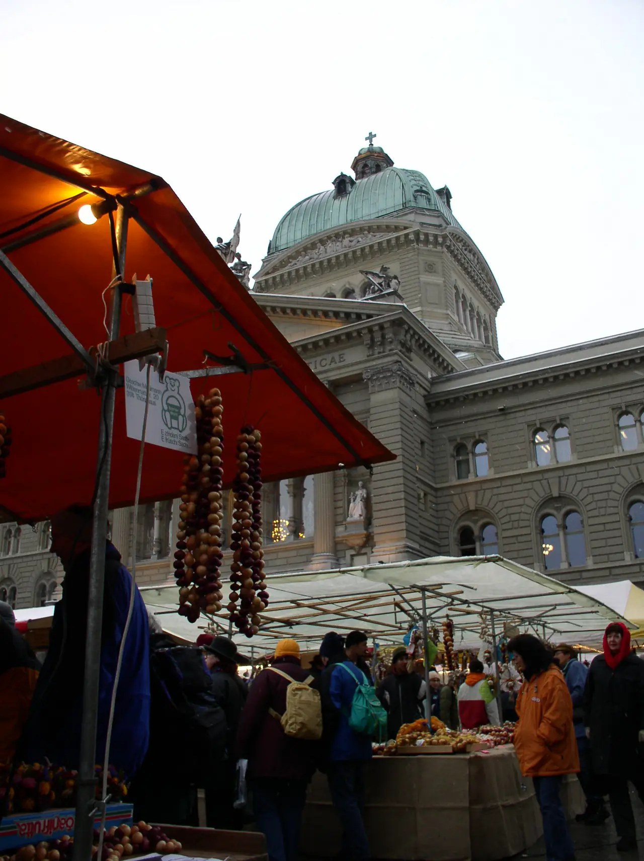 แผงขายของที่ตลาดหัวหอมในเบิร์น มีอาคาร Bundeshaus เป็นฉากหลัง ภาพจาก Wikimedia Commons ผู้ถ่าย David Buchmann สัญญา CC BY-SA 3.0 ไม่ทราบปีที่ถ่าย