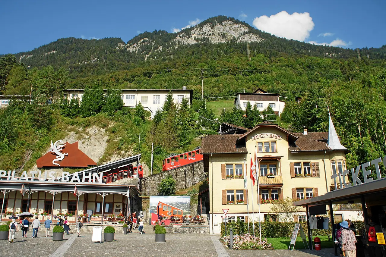 สถานีล่างของรถรางฟันเฟืองที่ Alpnachstad ป้าย Pilatus-Bahn และอาคารตั๋ว ภาพจาก Wikimedia Commons ผู้ถ่าย Mboesch สัญญา CC BY-SA 4.0 ไม่ทราบปีที่ถ่าย ฉากหลังเต็มไปด้วยต้นไม้ในฤดูร้อน