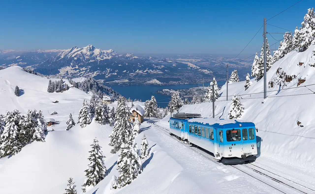 รถไฟของสาย Rigi (รุ่น BDhe 2/4 II ตามชื่อไฟล์) วิ่งบนทางที่มีหิมะระหว่าง Rigi Staffel กับ Rigi Kulm ฉากหลังเห็นทะเลสาบและภูเขา ภาพจาก Wikimedia Commons ผู้ถ่าย Kabelleger และ David Gubler สัญญา CC BY-SA 4.0 ไม่ทราบปีที่ถ่าย เป็นภาพฤดูหนาว