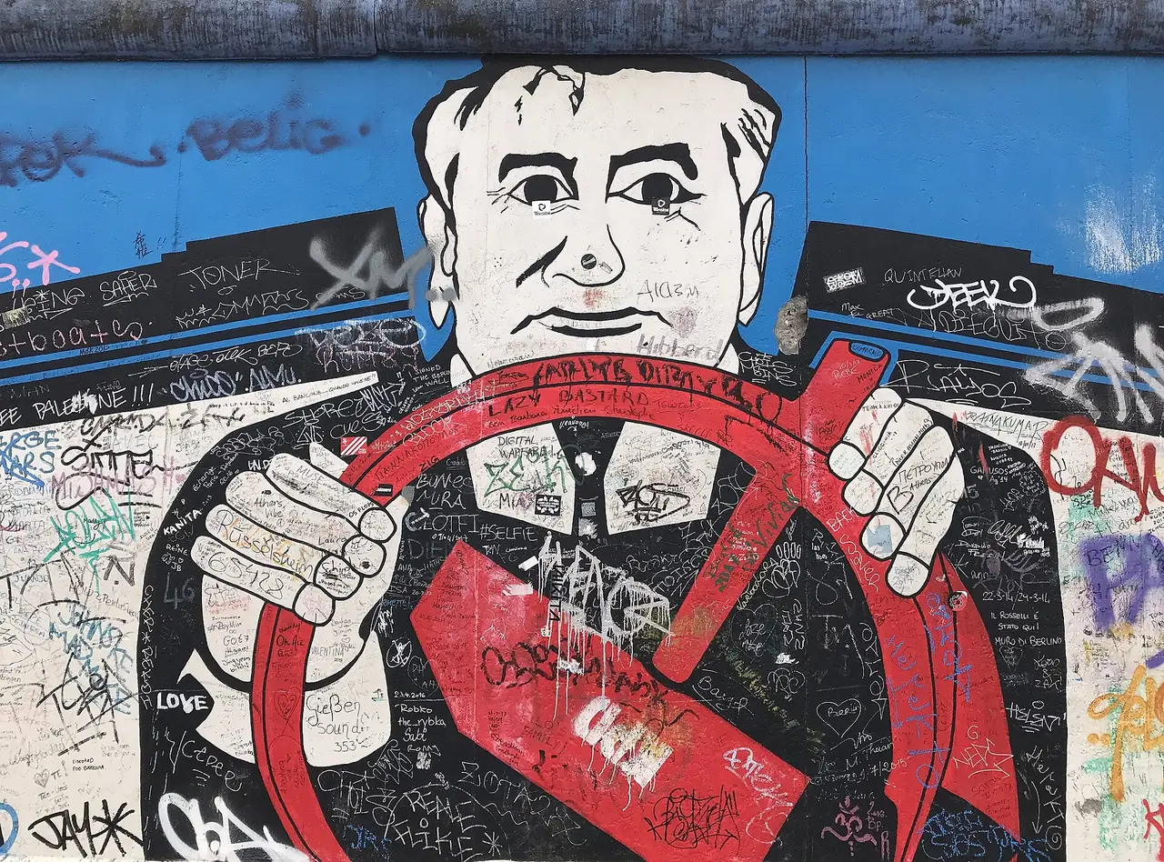 ภาพวาดบนกำแพง East Side Gallery รูปชายถือพวงมาลัย ผิวภาพมีลายเซ็นกราฟฟิตีของผู้มาเยือนทับอยู่ ภาพจาก Wikimedia Commons ผู้ถ่าย Dudva สัญญา CC BY-SA 4.0 ปีที่ถ่ายไม่ระบุ