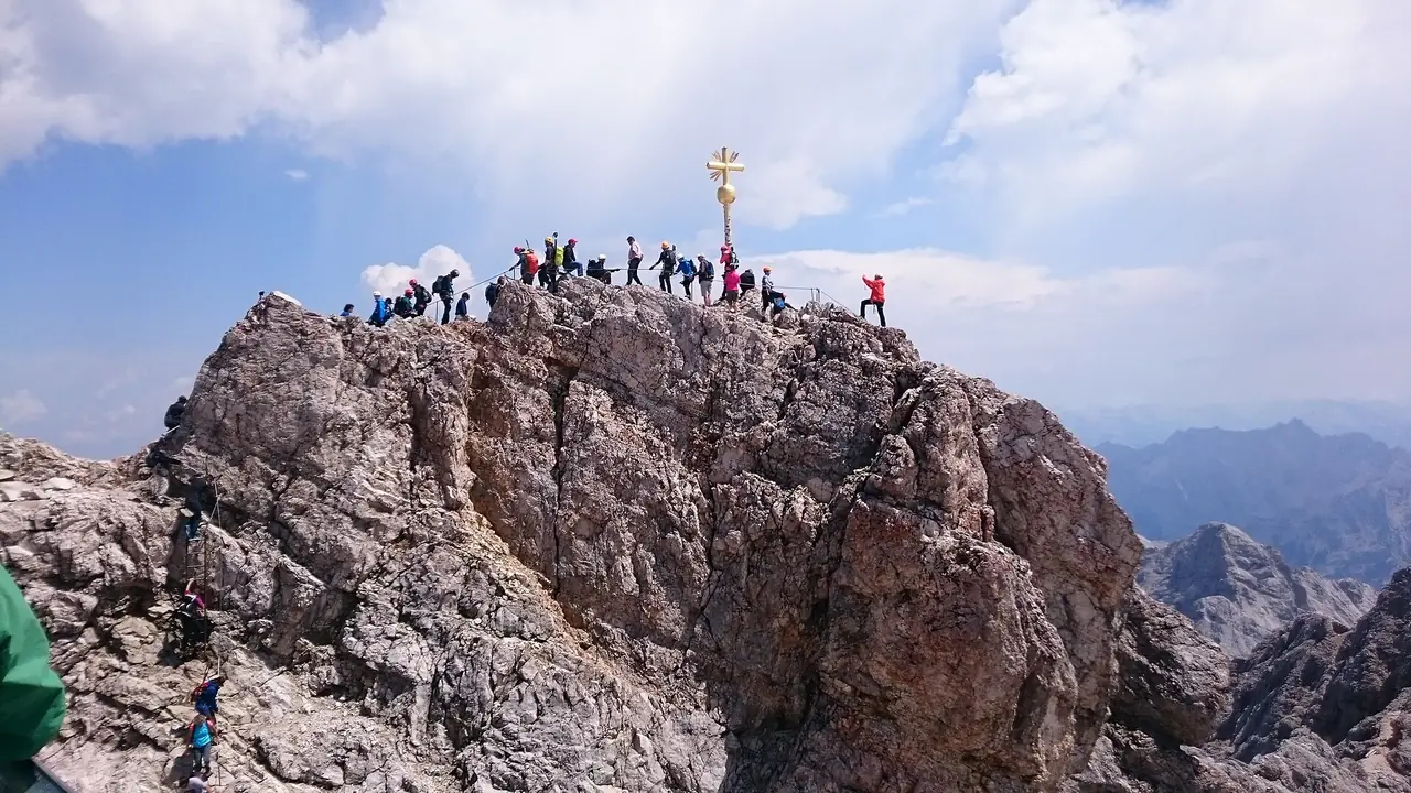 นักปีนเขาบนยอดหินใกล้ไม้กางเขนทองบน Zugspitze ภาพจาก Wikimedia Commons ผู้ถ่าย Treeem สัญญา CC BY-SA 4.0 ปีที่ถ่ายไม่ระบุ ภาพนี้เป็นส่วนหินที่ปีนไปถึง ไม่ใช่ลานสถานีที่คนที่ขึ้นกระเช้าเดินไปถึงโดยทั่วไป