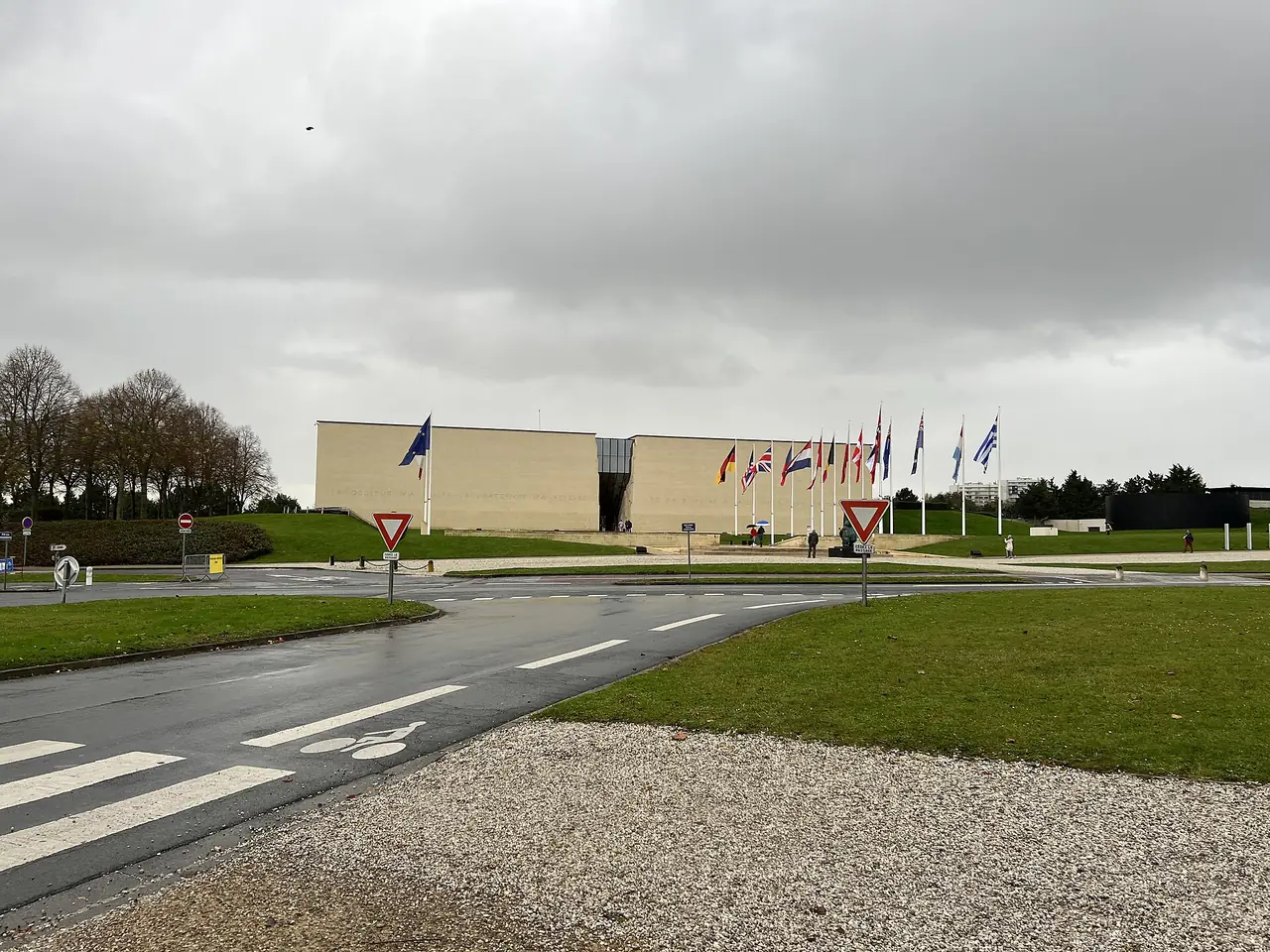 อาคารและเสาธงนานาชาติหน้า Mémorial de Caen ในวันฟ้าครึ้มและถนนเปียก ภาพถ่ายปี 2021 จาก Wikimedia Commons ผู้ถ่าย Chabe01 สัญญา CC BY-SA 4.0