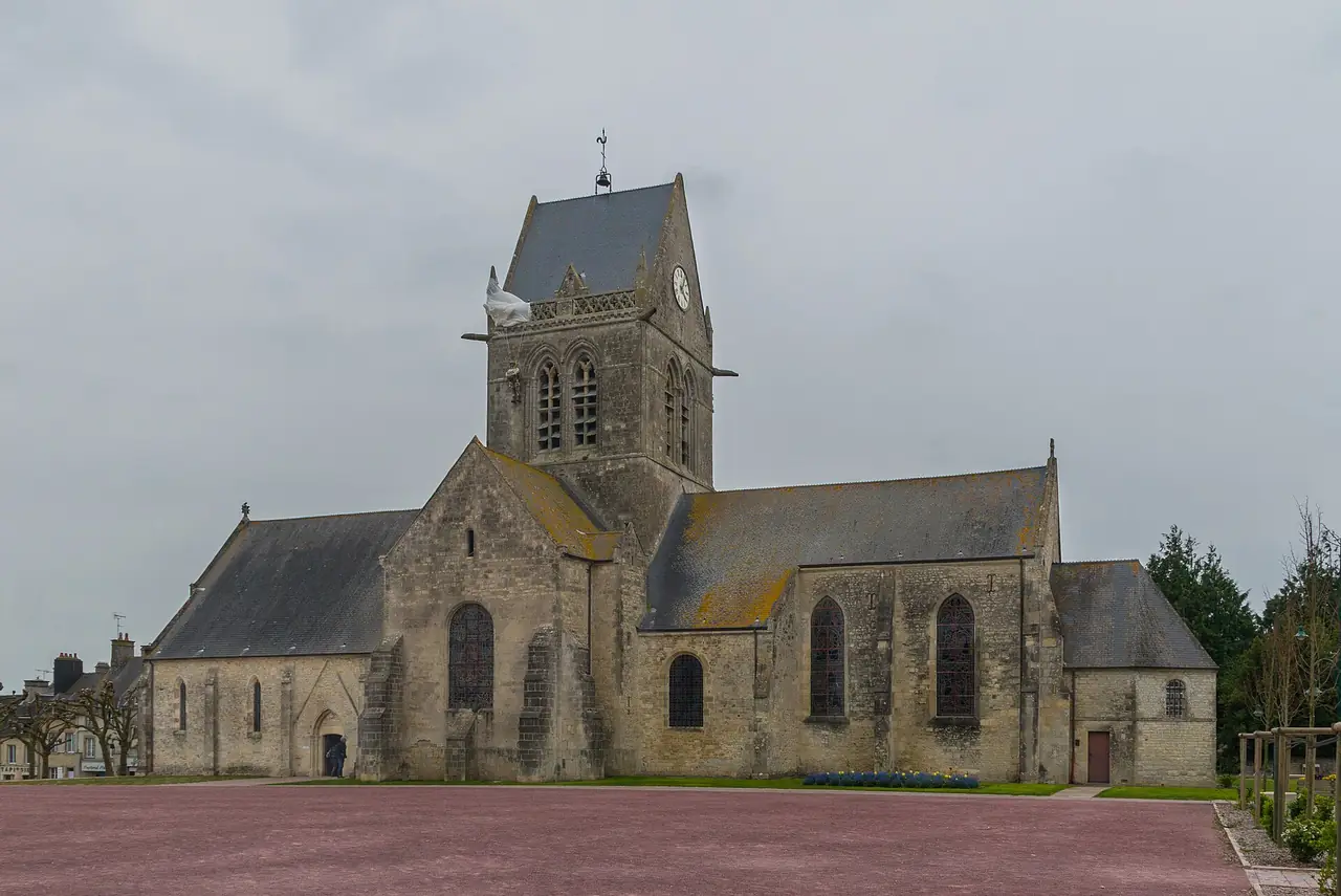 โบสถ์ Sainte-Mère-Église มองจากลานหน้าโบสถ์ มีหุ่นทหารพลร่มแขวนที่หอโบสถ์ตามเรื่องที่เล่าสืบมา ภาพจาก Wikimedia Commons ผู้ถ่าย Jebulon สัญญา CC0