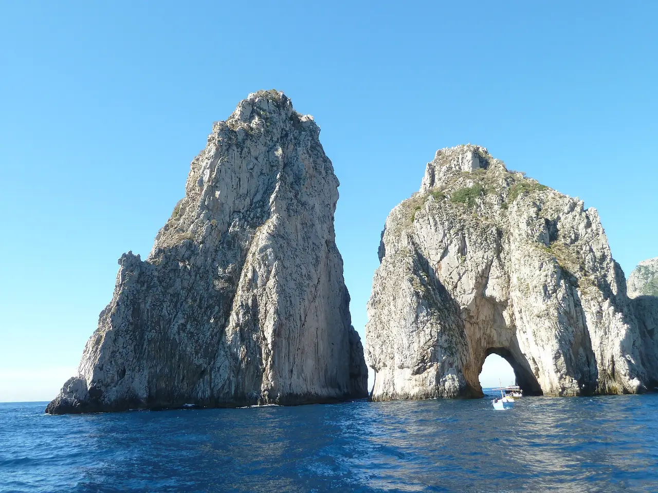 หินสูง Faraglioni นอกชายฝั่งคาปรี มองจากเรือ มีเรือลำเล็กกลอนผ่านช่องหิน ภาพจาก Wikimedia Commons ผู้ถ่าย Abxbay สัญญา CC BY-SA 4.0
